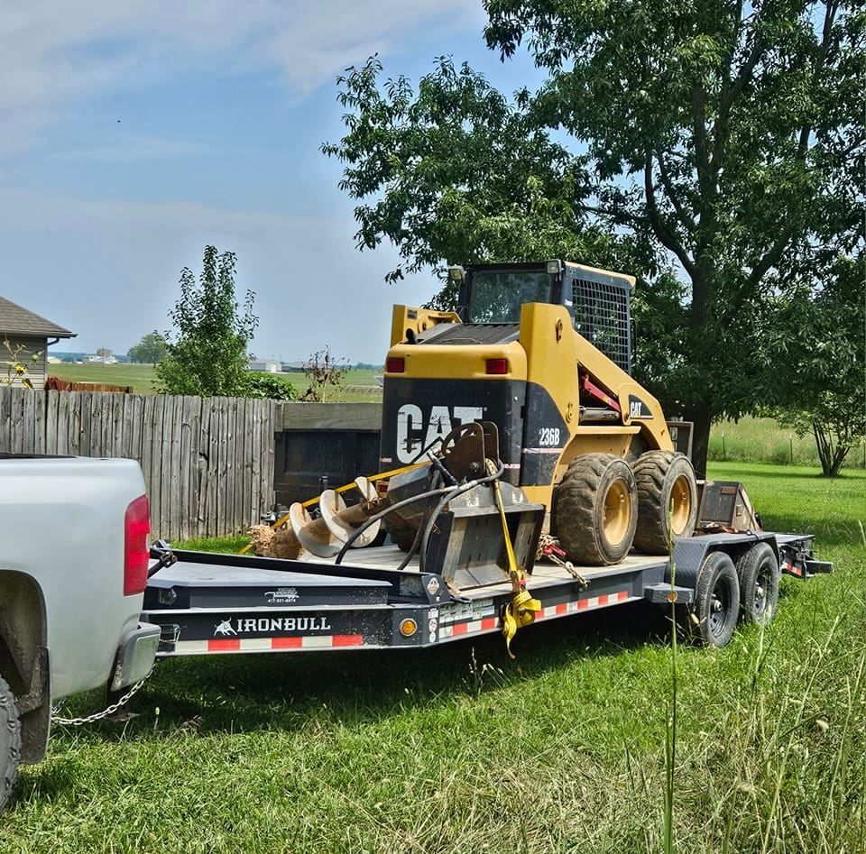 Yellow CAT skid steer on a trailer, attached to a white pickup truck, on grass near a fence.