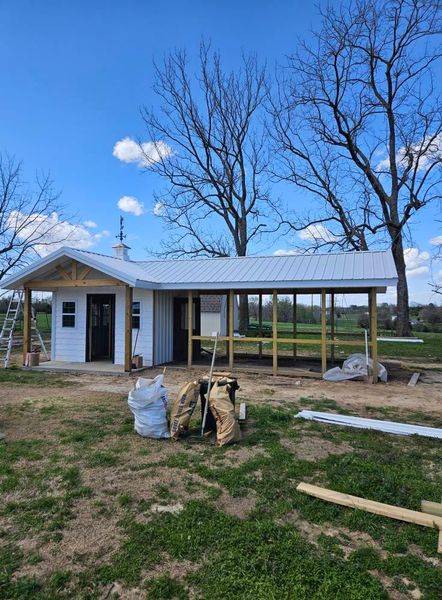 Building in progress; white building with porch and roof, construction materials in front, trees in the background.