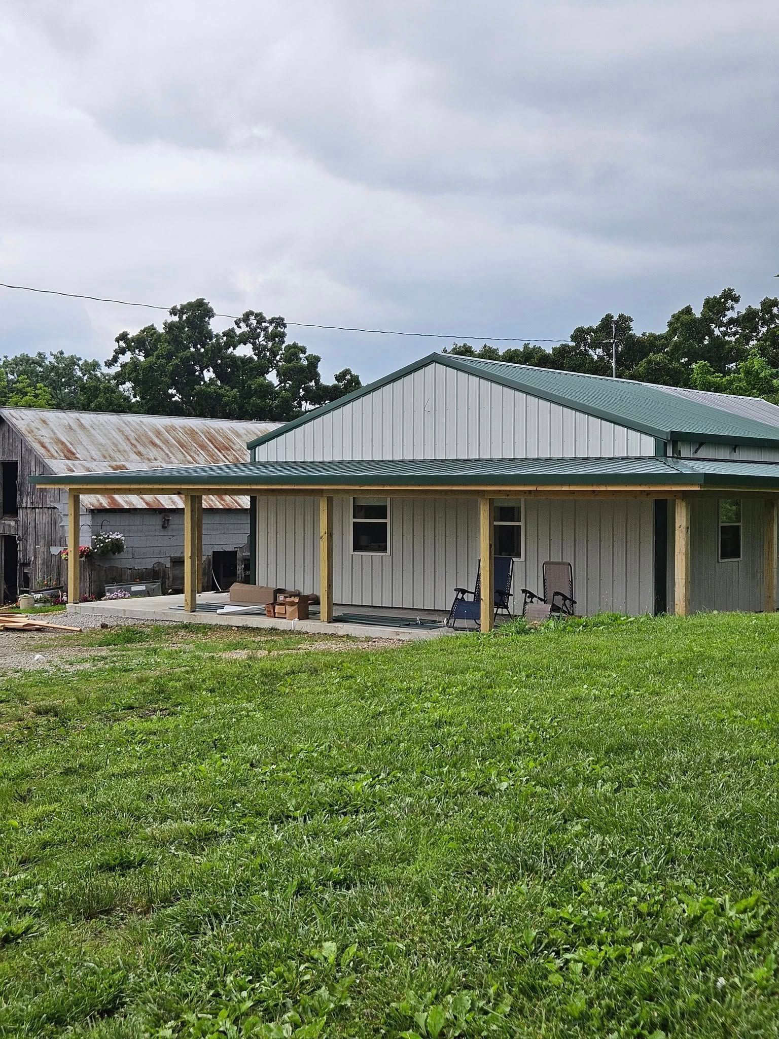 A newly built home with a green metal roof, covered porch, and white vertical siding sits next to a weathered barn.