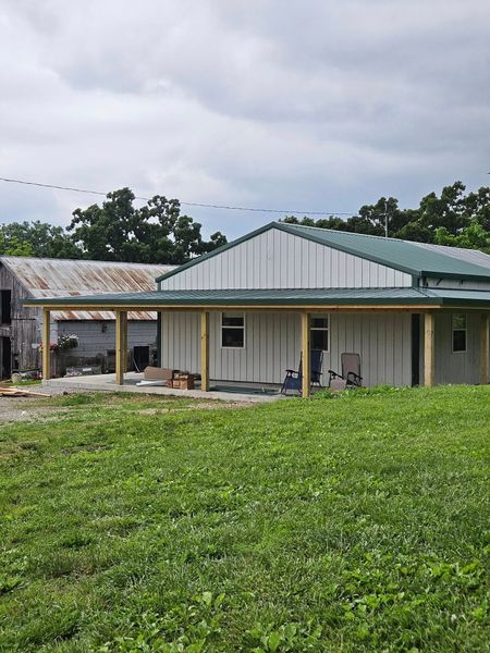 A newly built home with a green metal roof, covered porch, and white vertical siding sits next to a weathered barn.