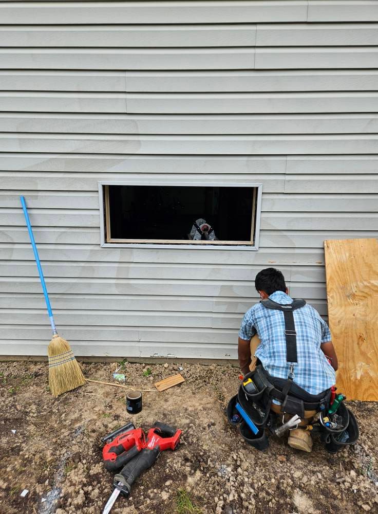 Two construction workers installing a window in a gray-sided building. One is inside, one outside, tools on the ground.