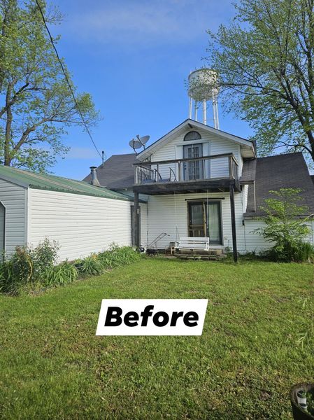 White house with balcony, water tower in background, and overgrown lawn.