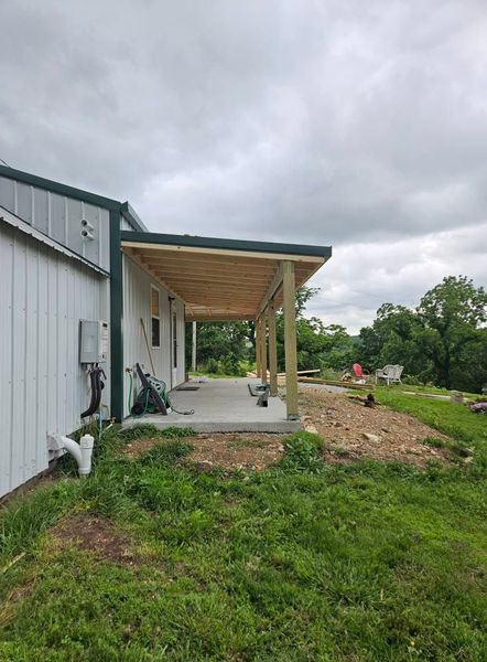 A new wooden patio cover extends from a white building with a green roof, set on a concrete slab.