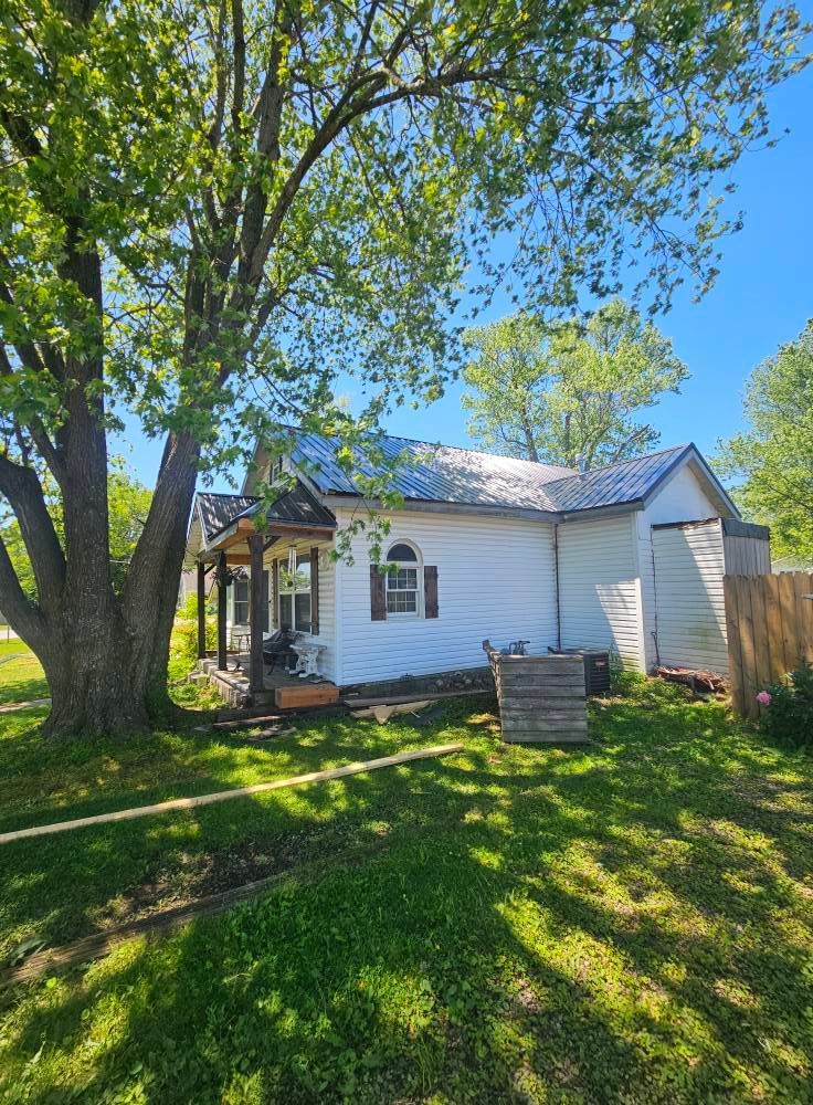 White house with metal roof, porch, and a large tree in front on a sunny day.
