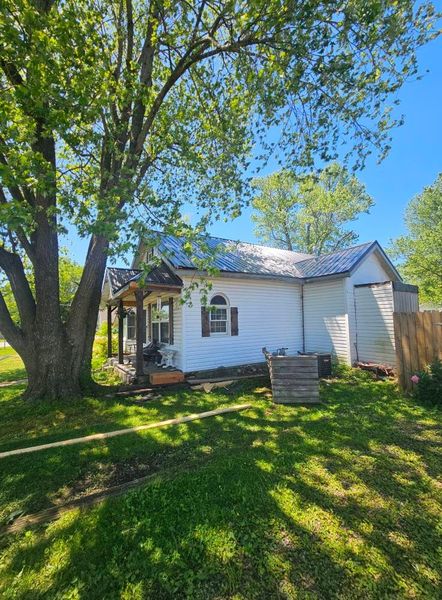 White house with metal roof, porch, and a large tree in front on a sunny day.