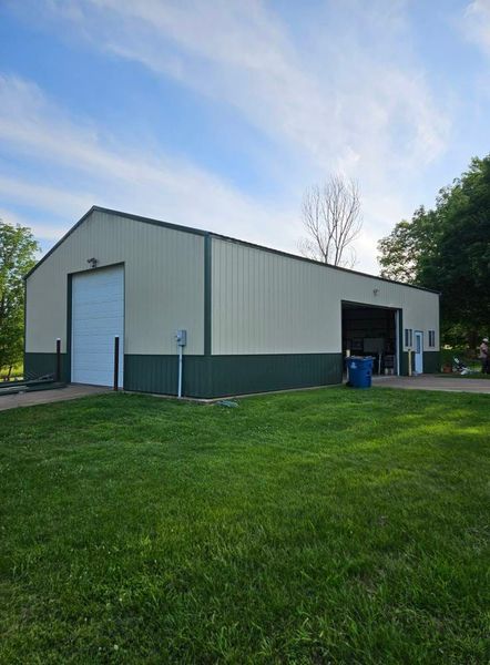 Metal building with tan and green siding, garage door, grassy lawn, and blue sky.