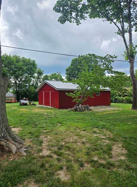 Red barn with a silver roof in a grassy yard, surrounded by trees under a cloudy sky.