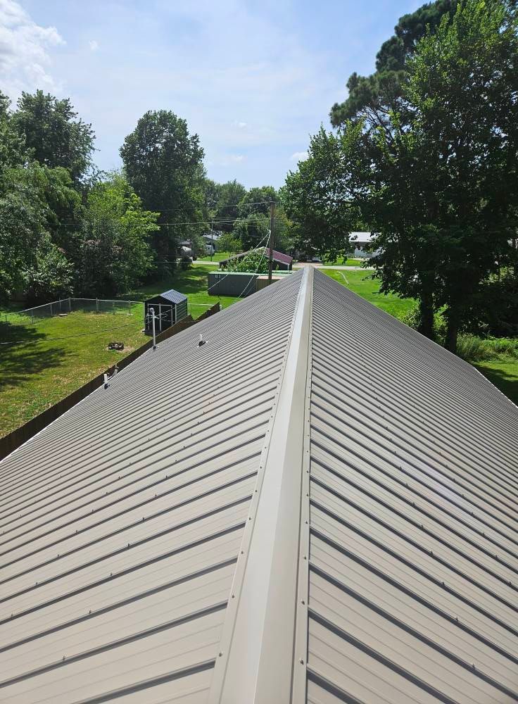 Metal roof of a building with visible ridges. Green trees and grass surround the structure.