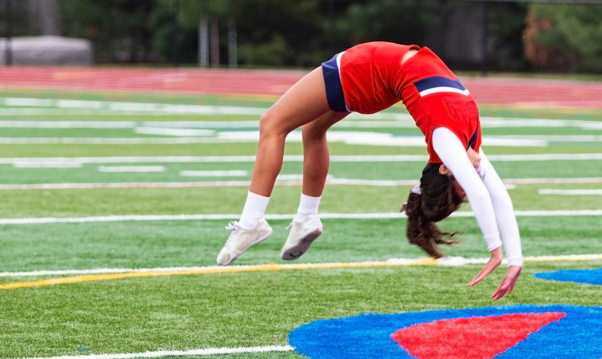 Cheerleader in red uniform performs a backbend on a green and red football field.