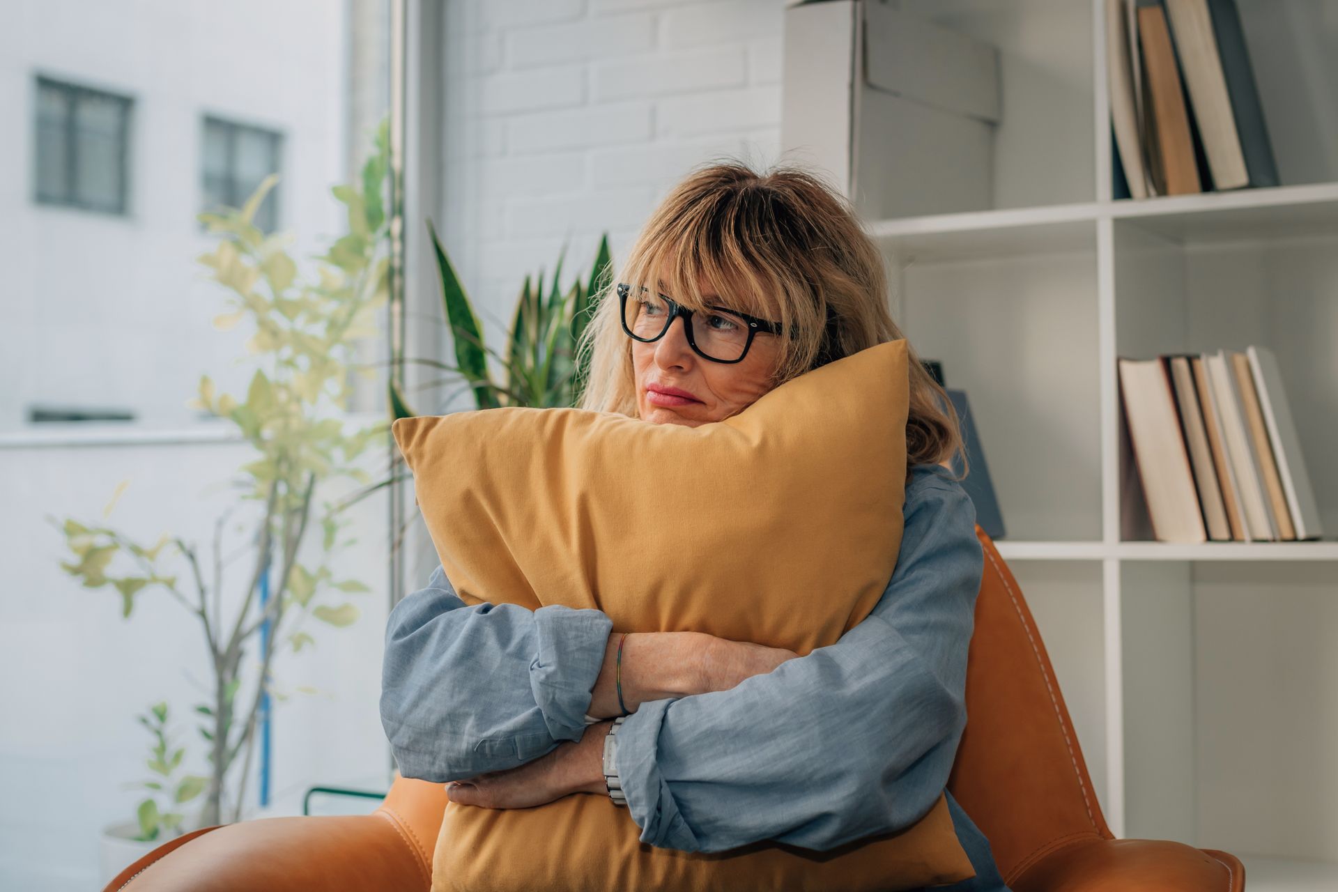 Person hugging a large orange pillow on a couch, looking thoughtful in a bright room