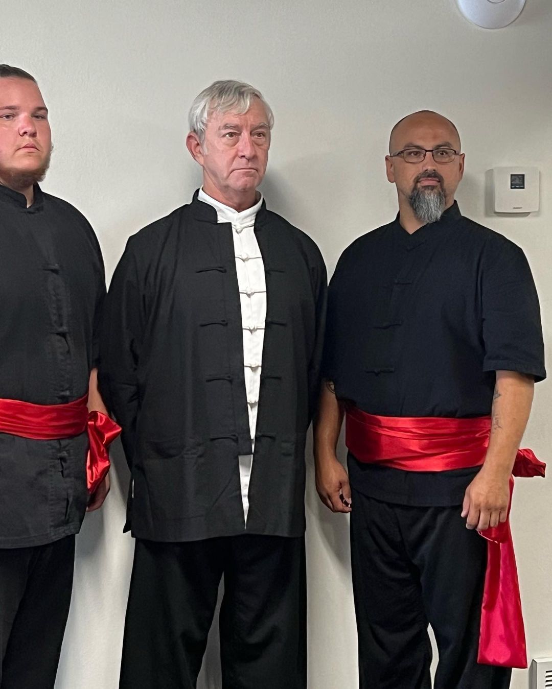 Three men are standing next to each other with red sashes around their waists