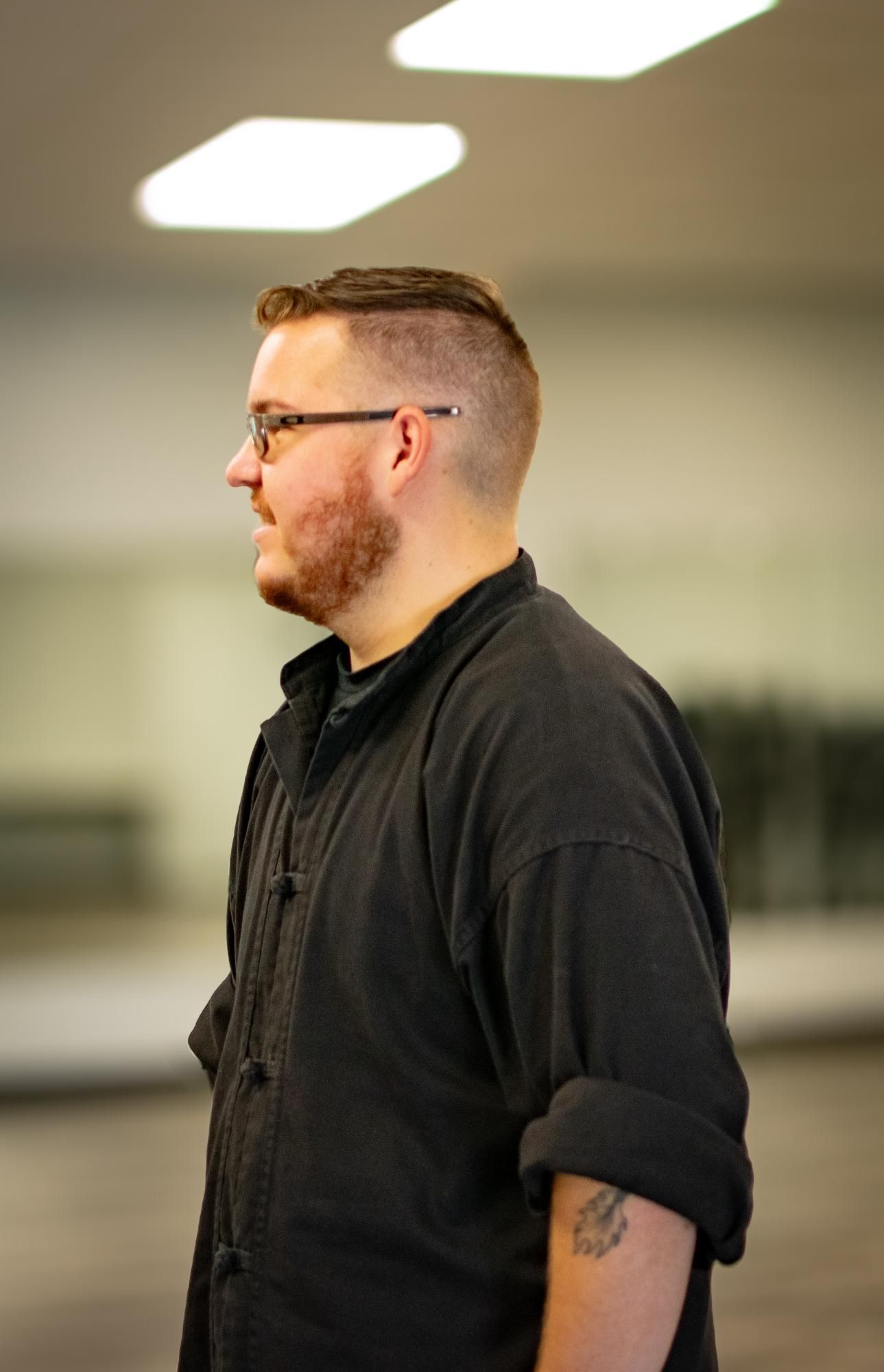 Man with glasses and beard, in a black outfit, standing in a well-lit room, looking to the left.