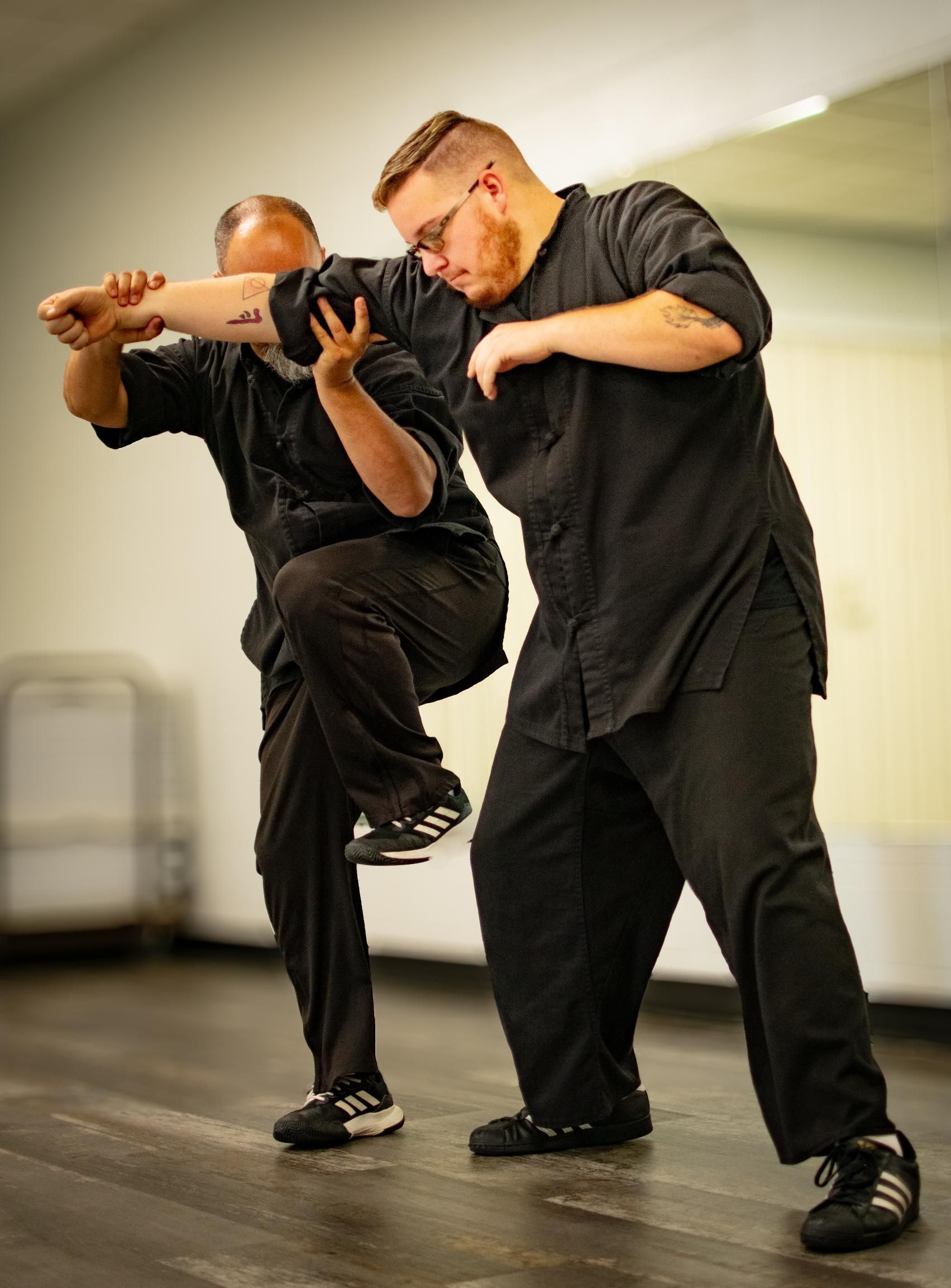 Two men in black martial arts uniforms practicing a self-defense technique indoors; one raises a knee while the other blocks.