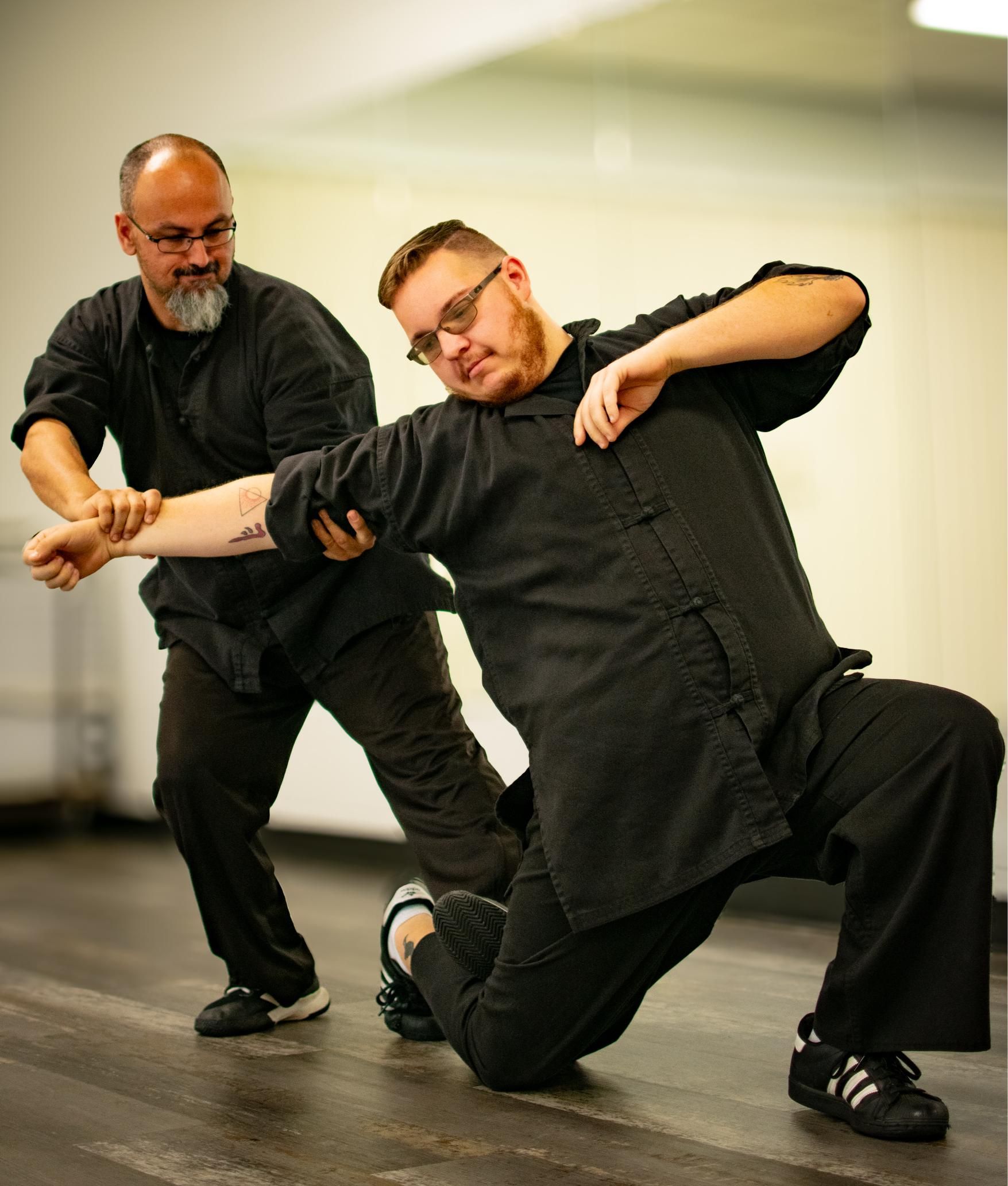Two men in black martial arts attire practice a self-defense move in a studio. One holds the other's arm.