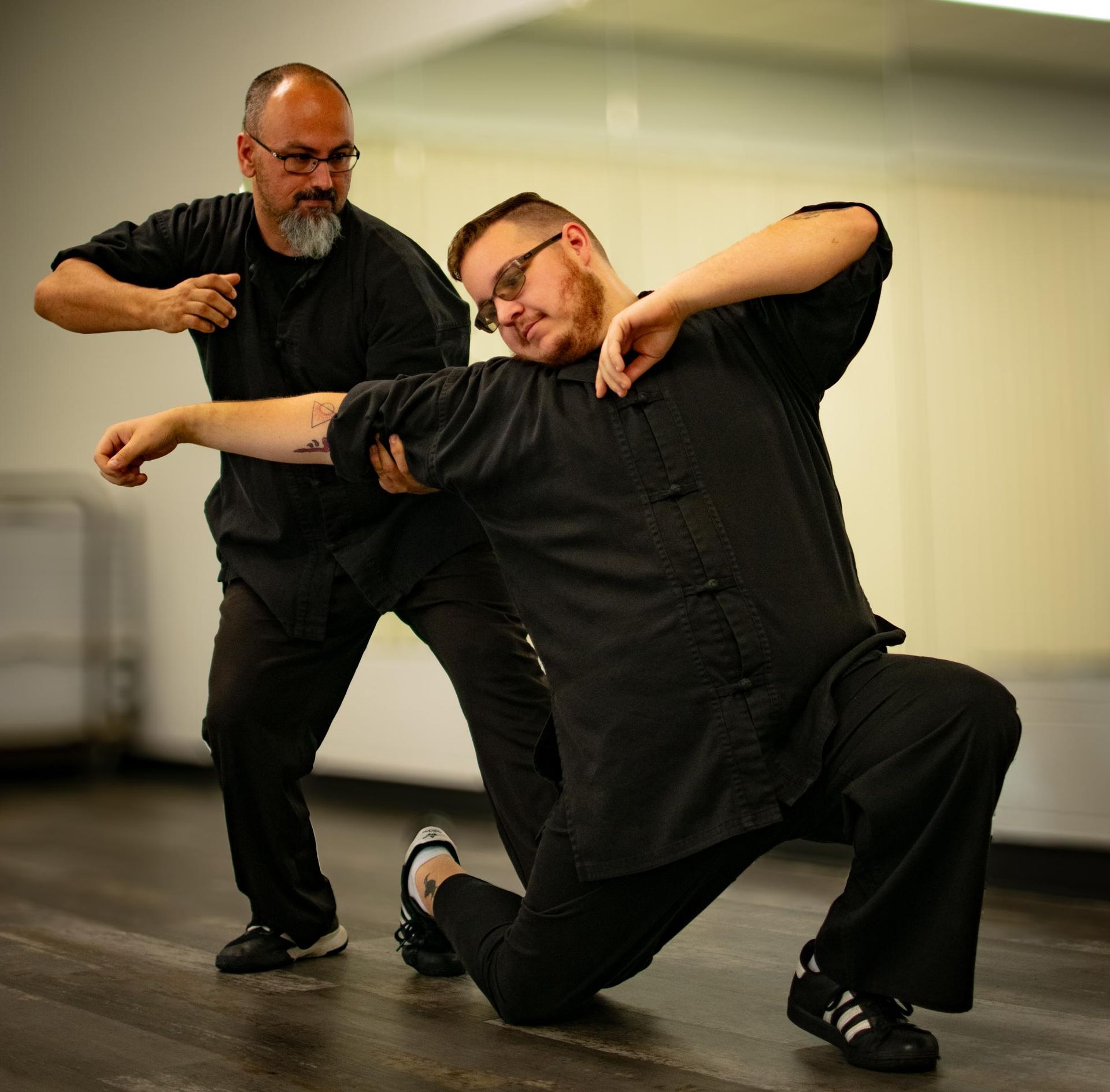 Two men in black uniforms practice martial arts in a room. One kneels, being blocked by the other.