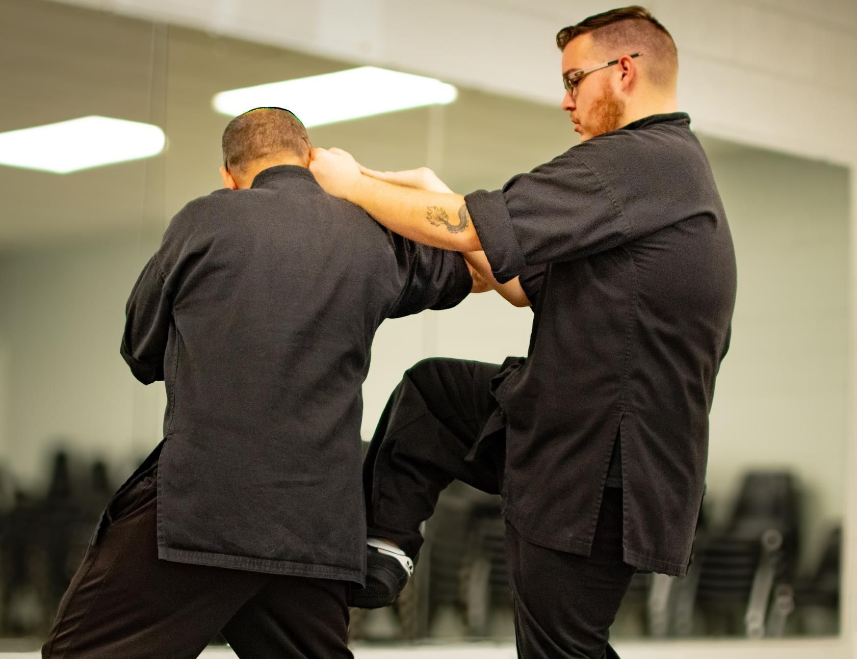 Two men in black practicing martial arts. One kicks while the other blocks and grabs. Indoors.