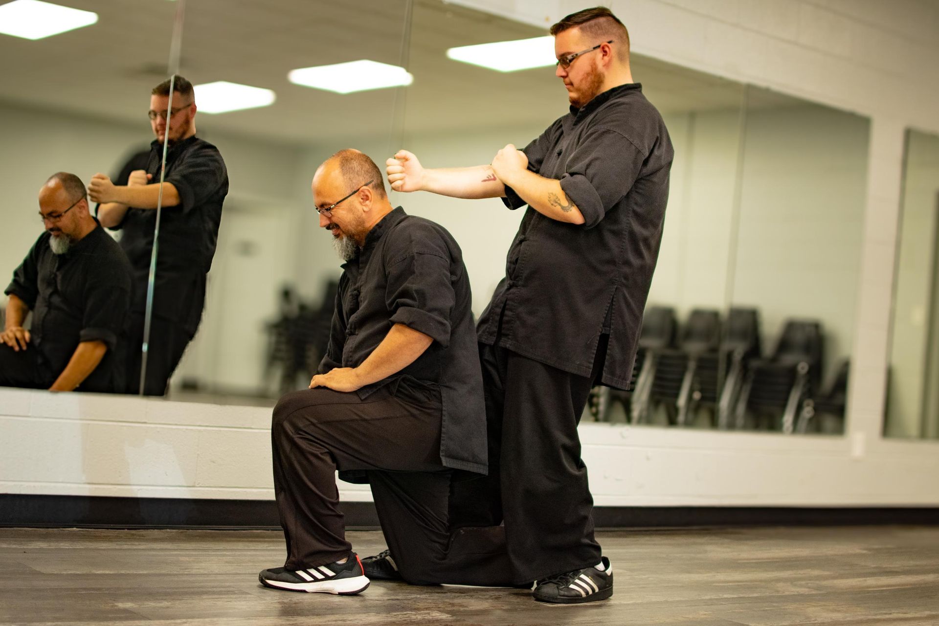 Two men in black practicing martial arts in a studio with mirrors. One kneels as the other demonstrates a block.