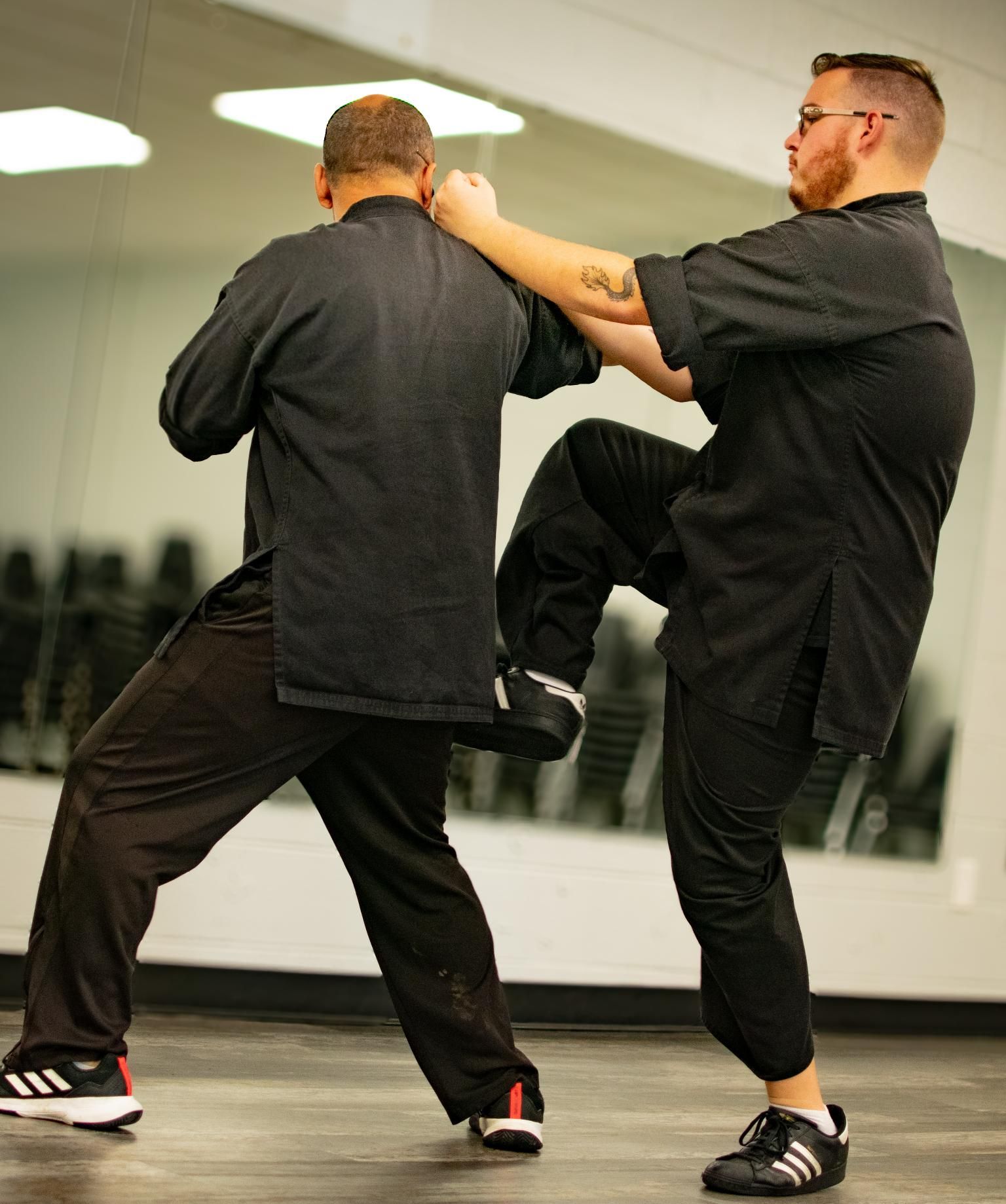Two men in black practicing martial arts. One kicks while the other blocks. In a studio.