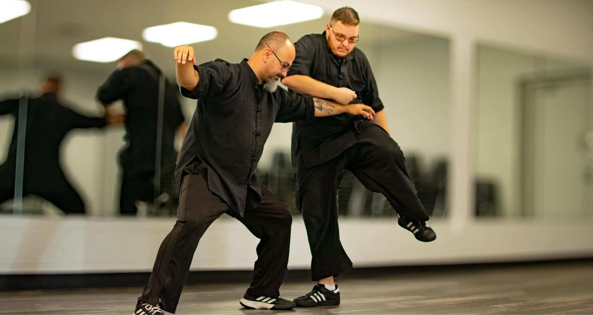 Two men in black martial arts uniforms practicing in a studio with mirrors. One kicks, the other blocks.