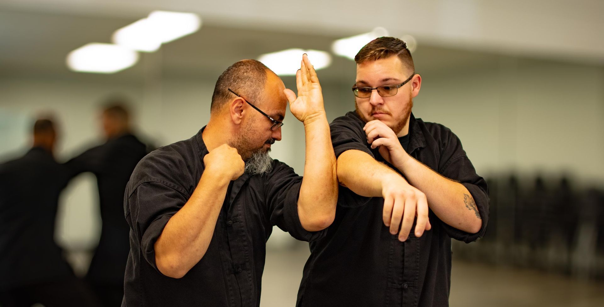 Two men in black shirts practicing martial arts in a studio. The man on the left blocks with his right hand.