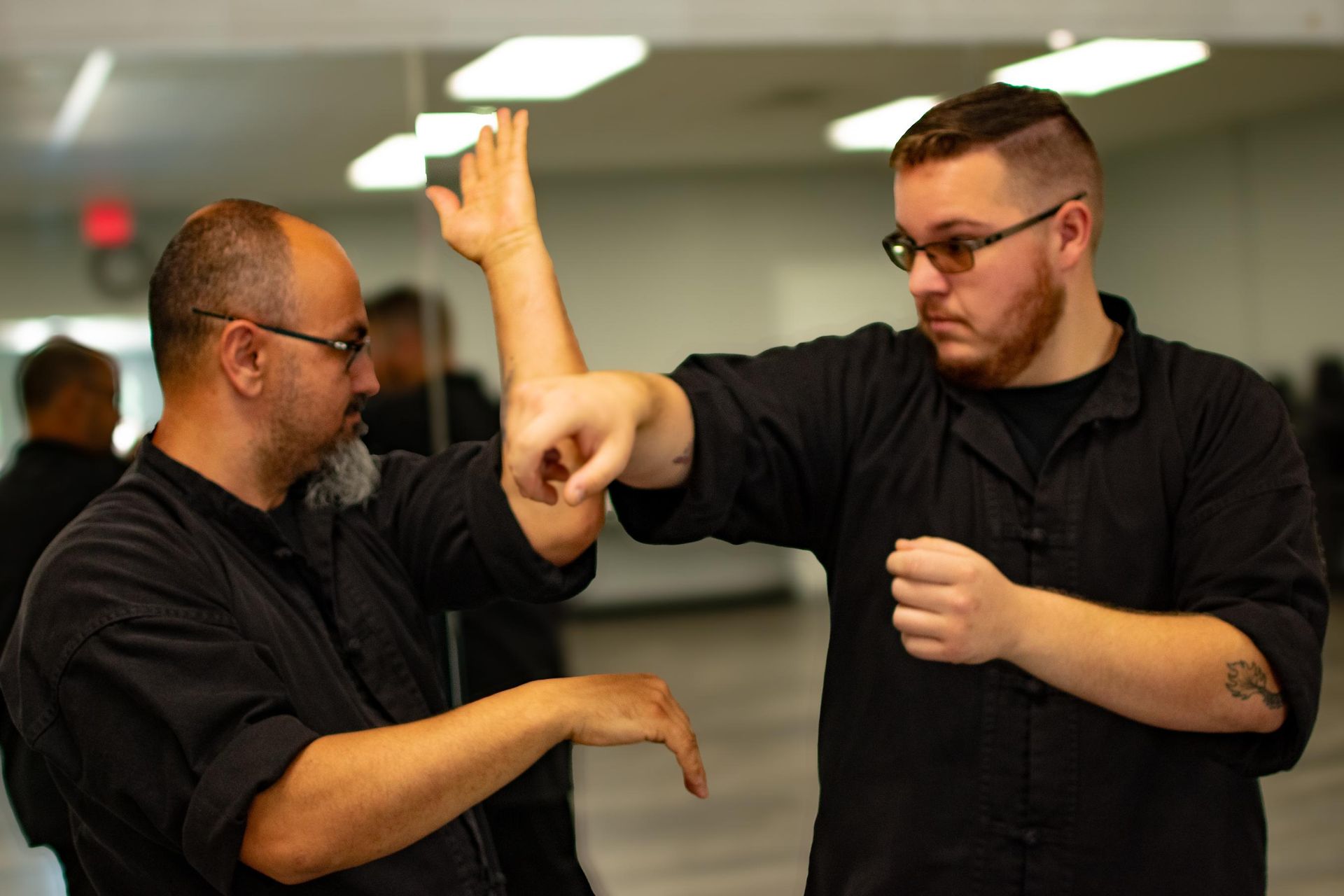 Two men in black shirts practice martial arts in a mirrored studio. One blocks a punch.