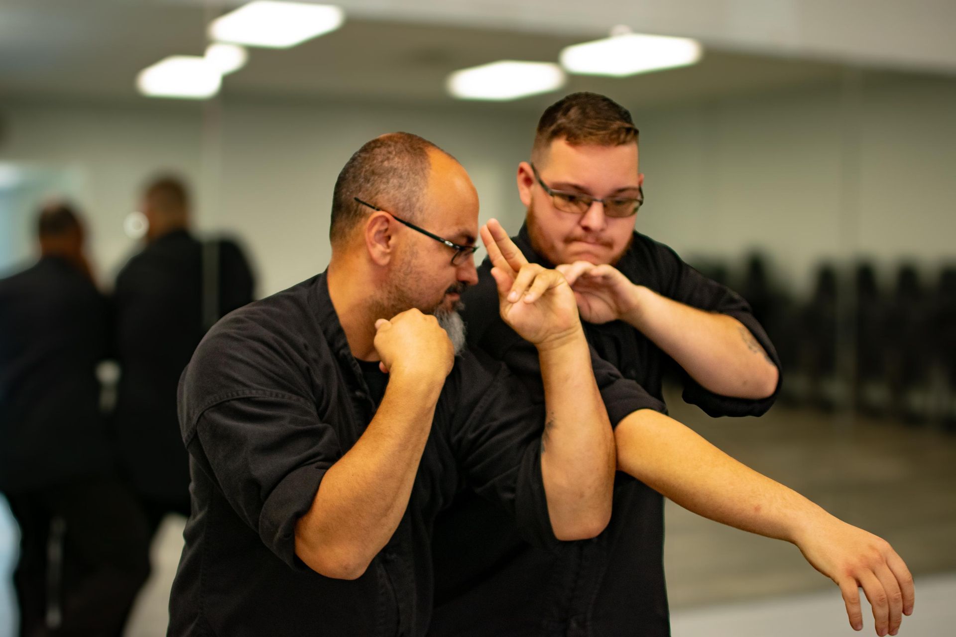 Two men in black shirts practice a martial arts move in a studio with mirrors.