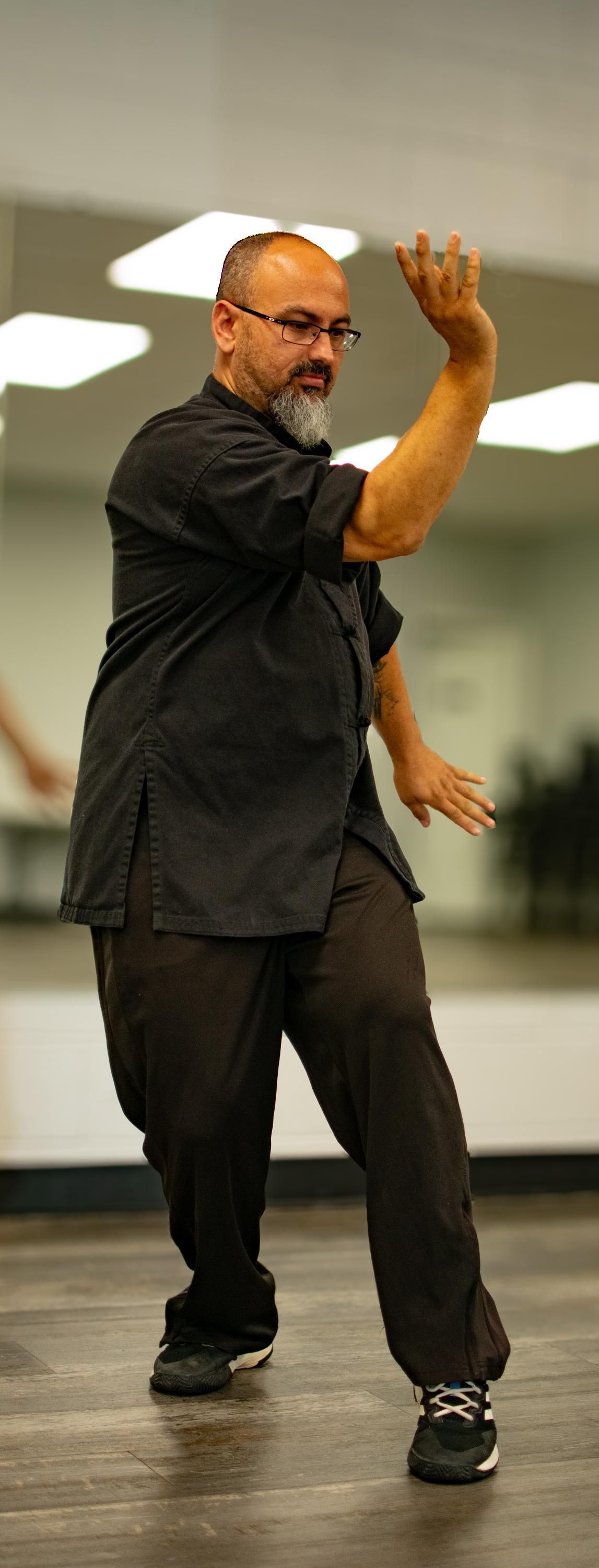 Man in black clothing practices tai chi. Indoors with mirrors, a wooden floor. He has glasses and a beard.