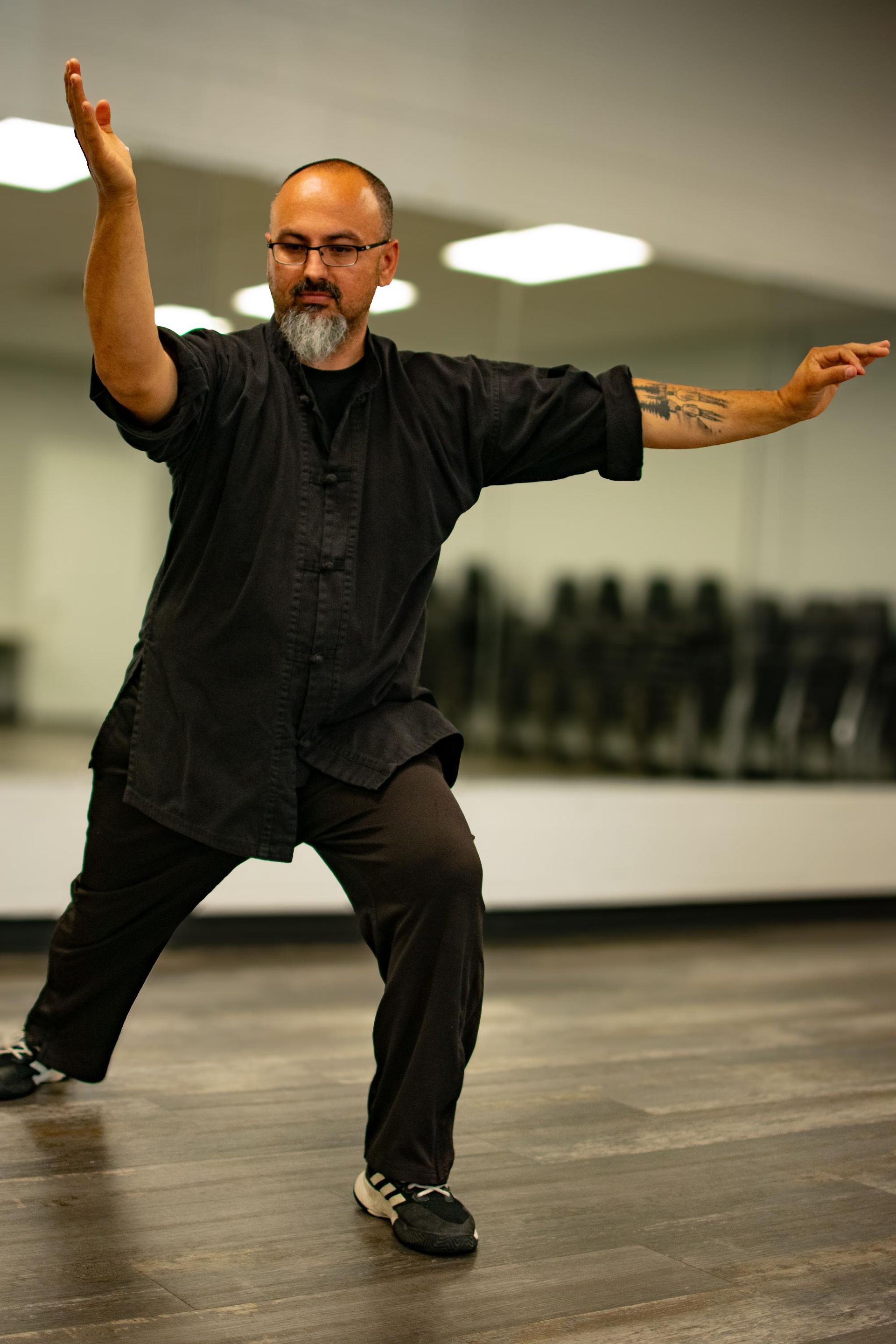Man in black martial arts attire, performing a Tai Chi pose in a studio with a mirror.