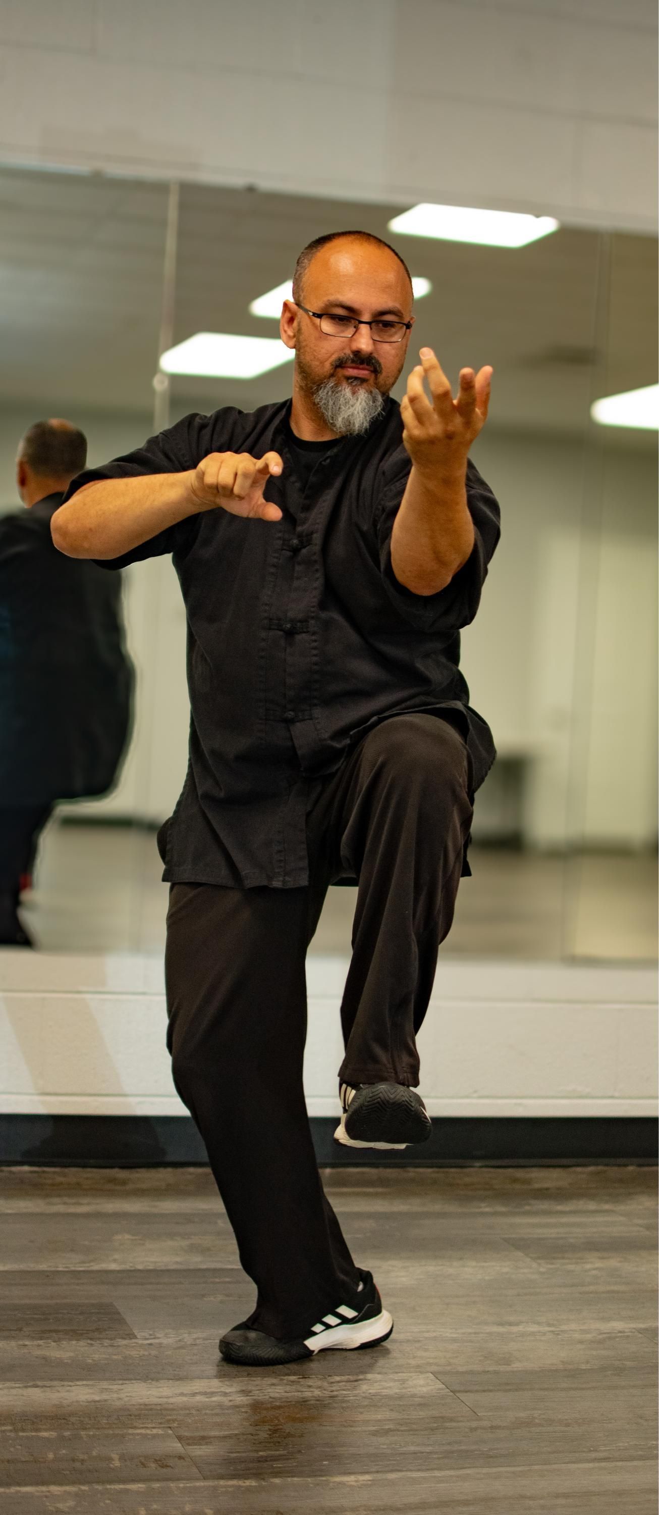 Man in black clothing performing a martial arts stance. He is indoors with a mirrored wall.