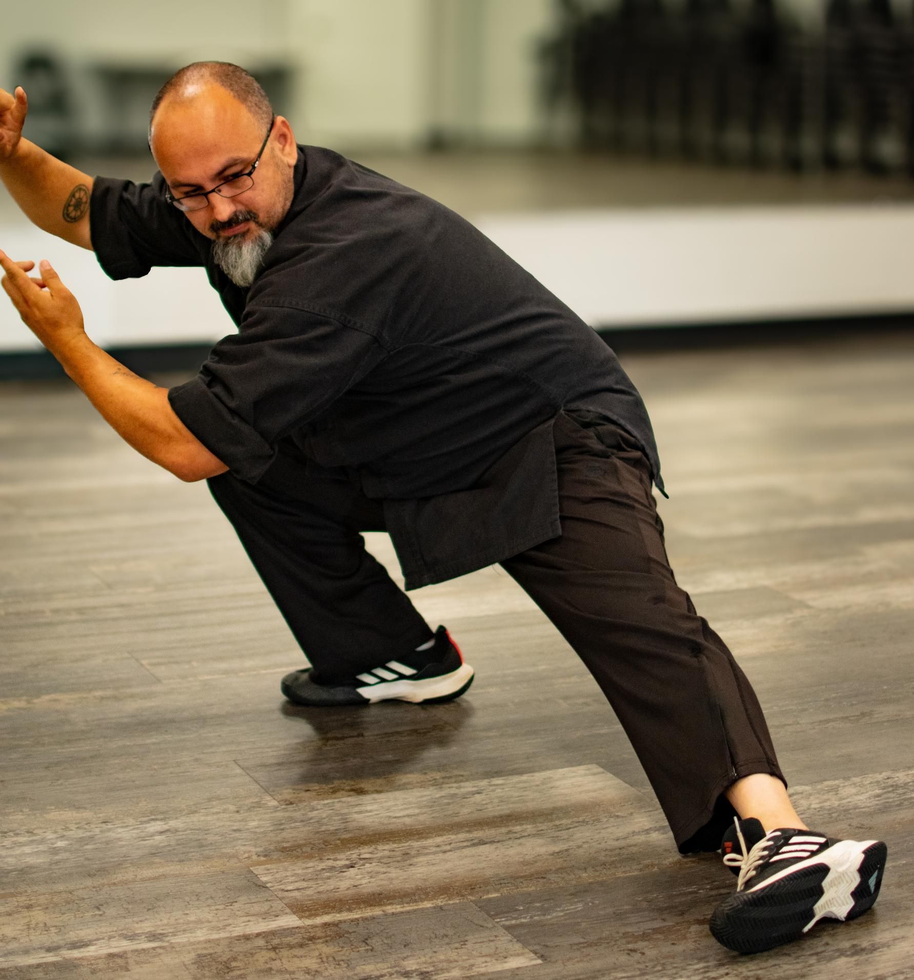 Man in black clothing practicing Tai Chi in a studio, demonstrating a low stance with focused expression.