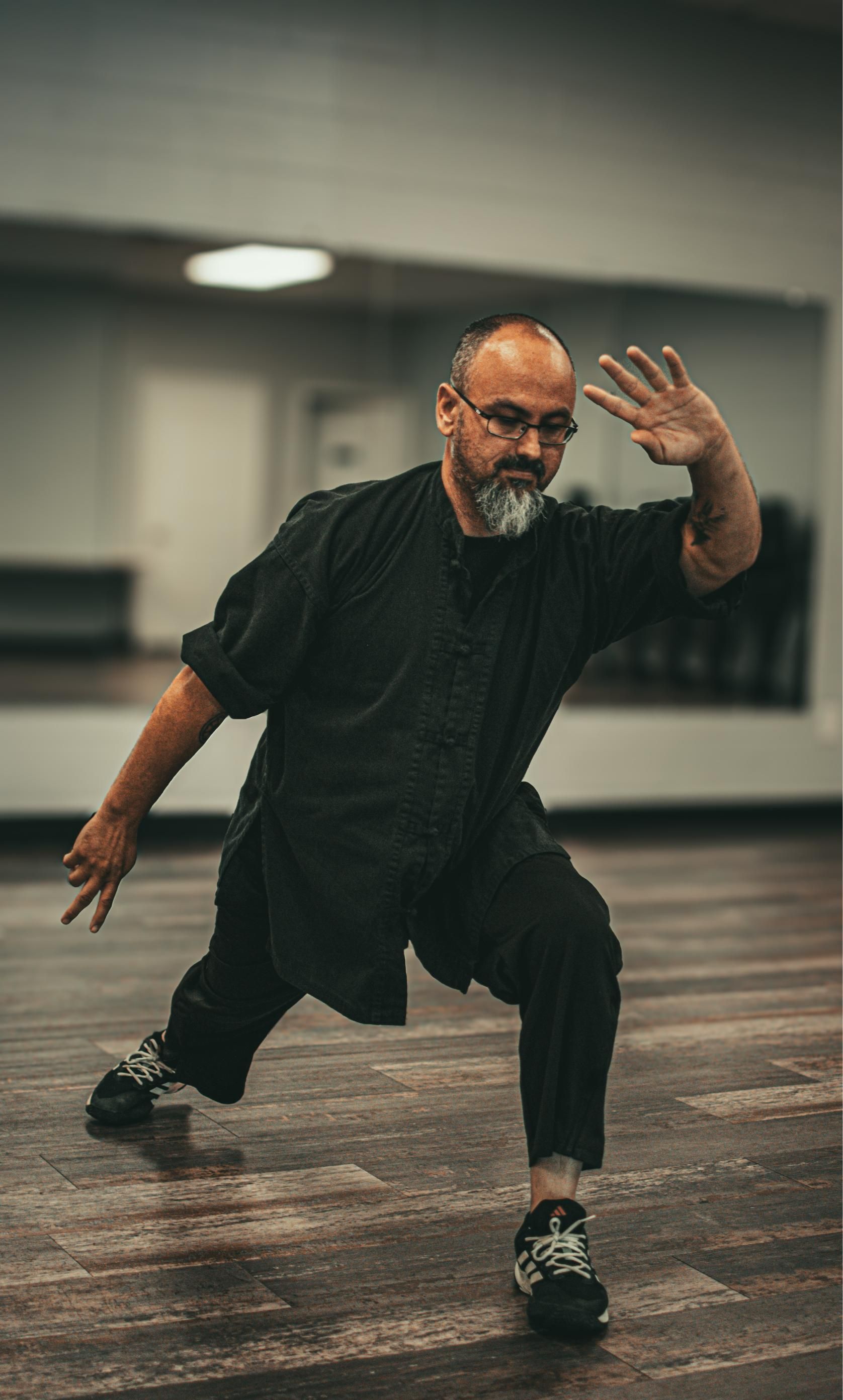 Man in black outfit practicing martial arts in a studio, with raised hand and squat.