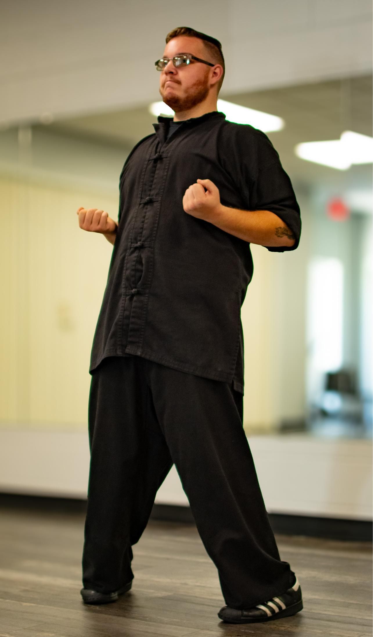 Man in black martial arts attire in a stance, fists clenched, in a dance studio.