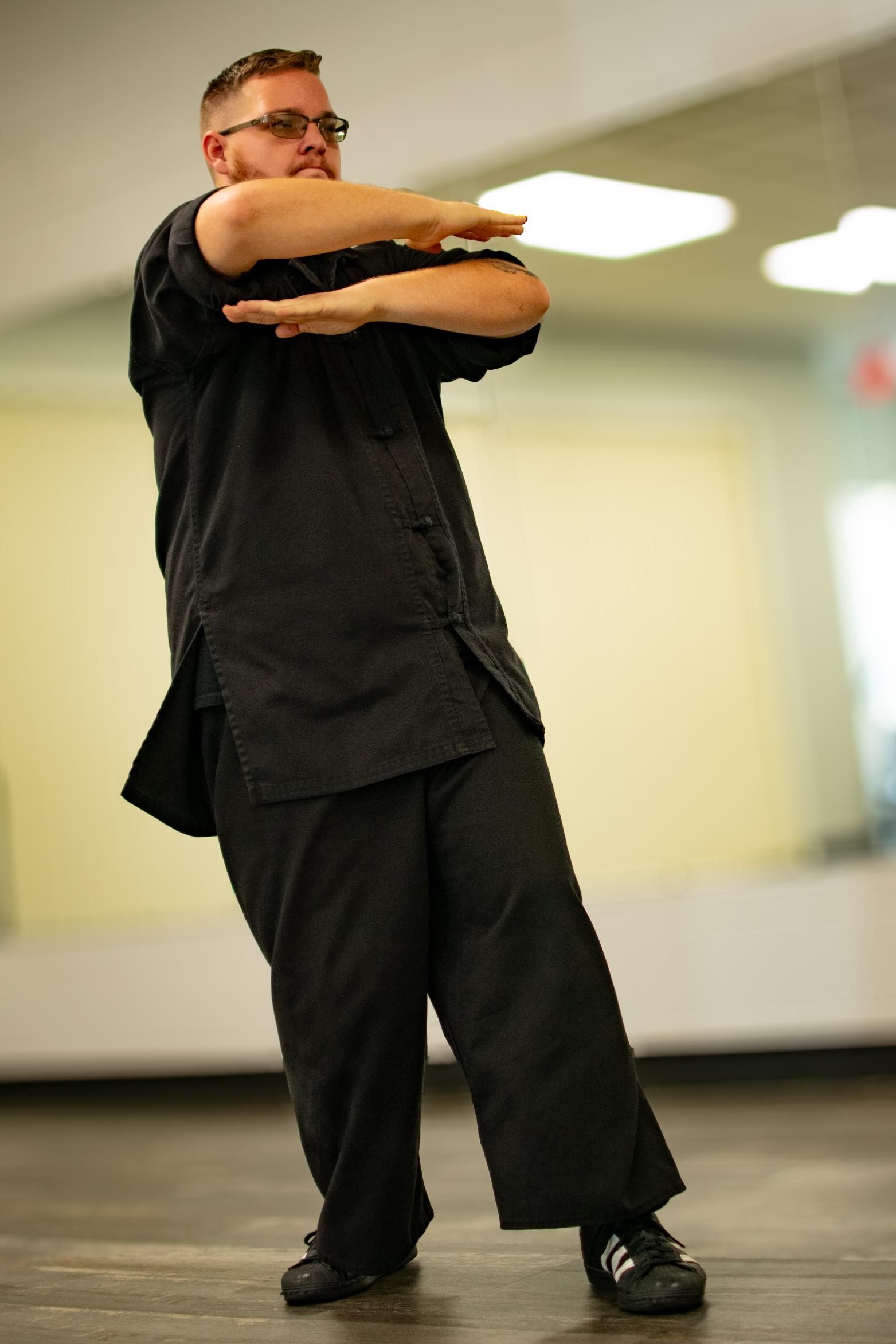 Man in black clothing performs a martial arts stance, arms crossed, in a studio.