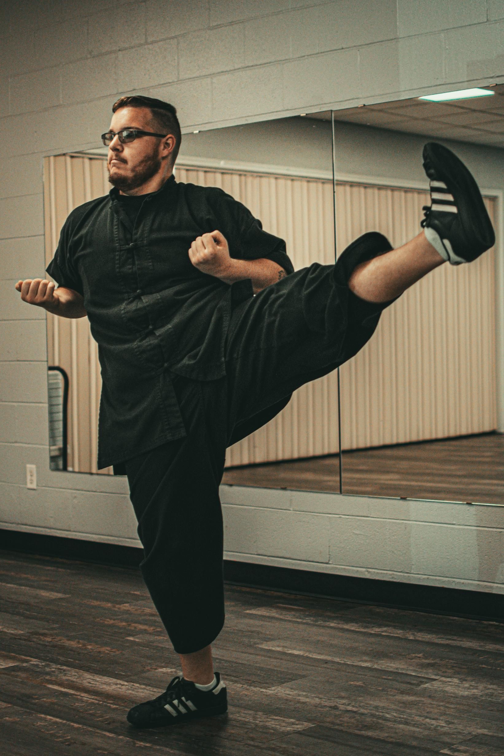 Man in black martial arts uniform, performing a high kick in a dance studio.