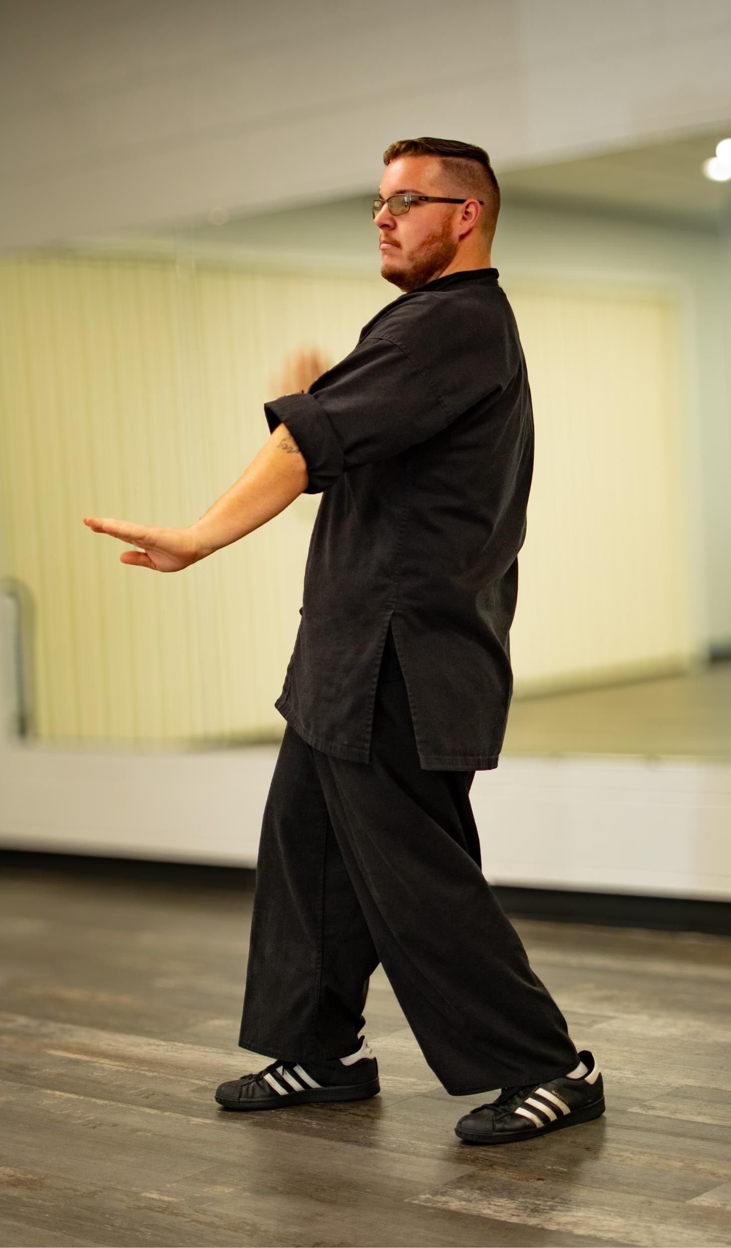 Man in black martial arts uniform, left arm extended, right fist up, practicing in a studio.