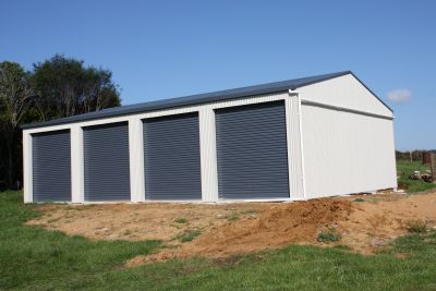 A white garage with three black roller doors in a grassy field.