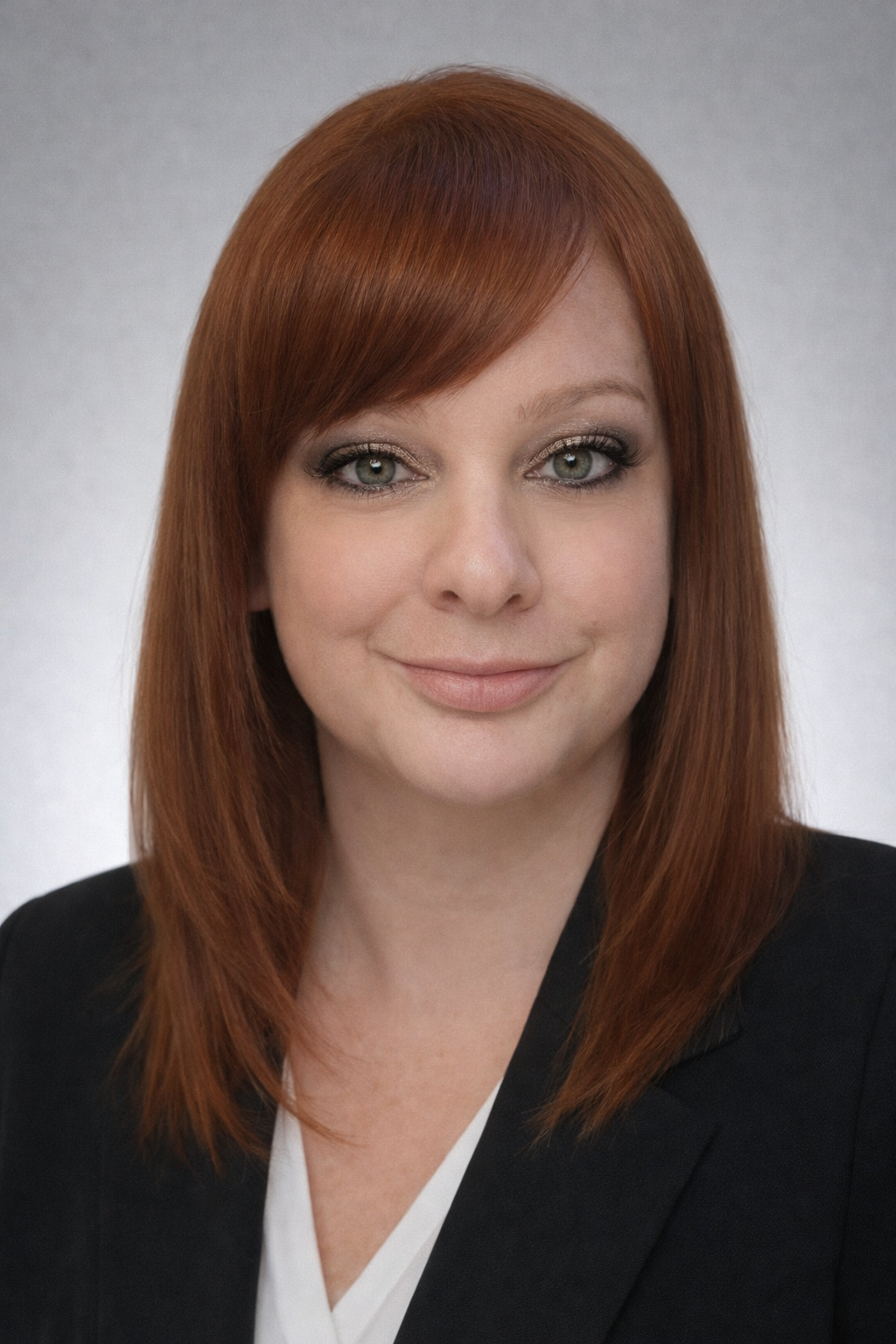 Woman with auburn hair and bangs, smiling, wearing a black blazer and white top, against a grey backdrop.