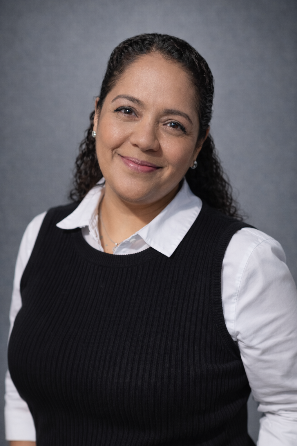 Woman with curly dark hair, wearing a white shirt and black sweater vest, smiles at the camera against a gray backdrop.