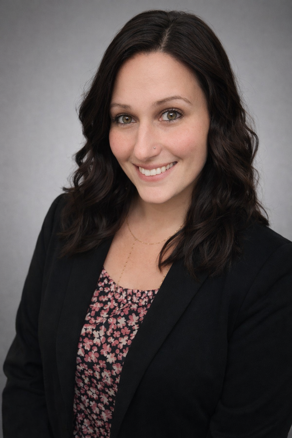 Woman with dark wavy hair smiles, wearing a black blazer over a floral print top, against a gray backdrop.