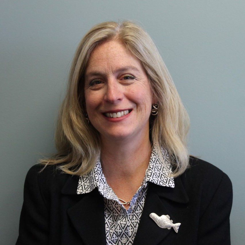 Lisa Bogan-Reohr wearing a patterned shirt and black blazer, smiling. A fish brooch is pinned on the blazer.