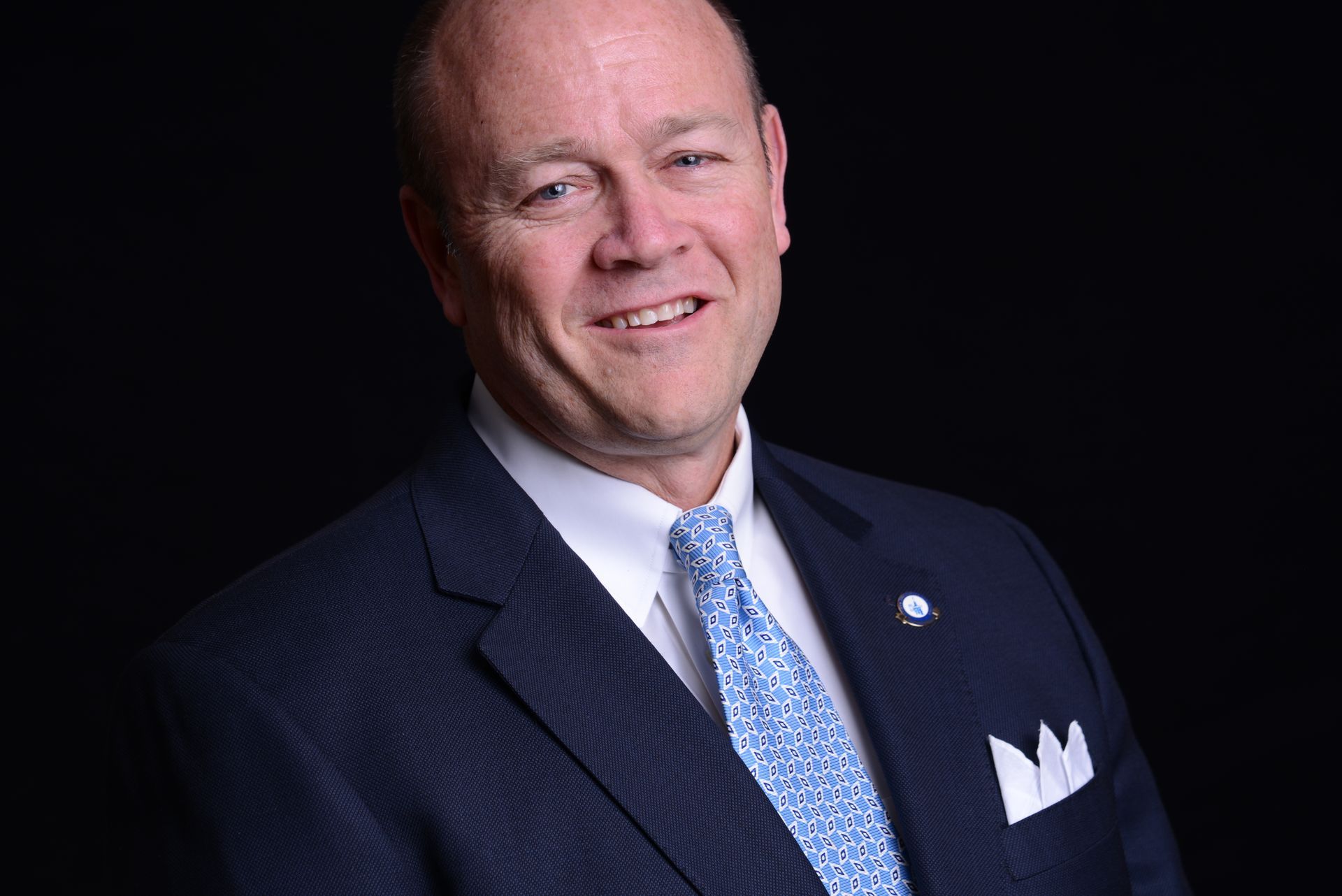 David V. Diggs in a blue suit, white shirt, and patterned tie smiles against a black backdrop.