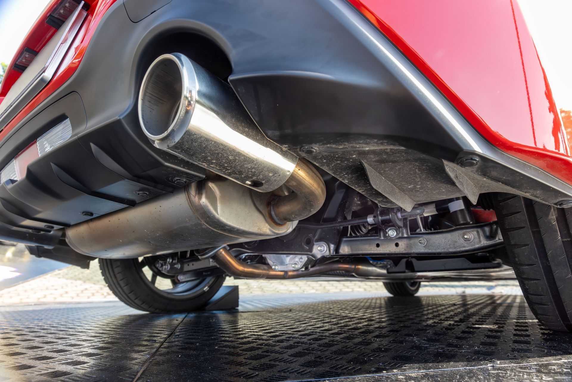 Red car's underside with exhaust pipe, showing muffler and rear tire on a textured surface.