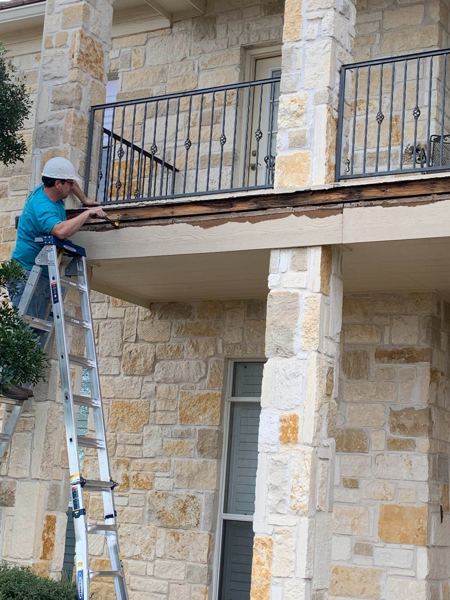 A man on a ladder is working on a balcony of a stone building.