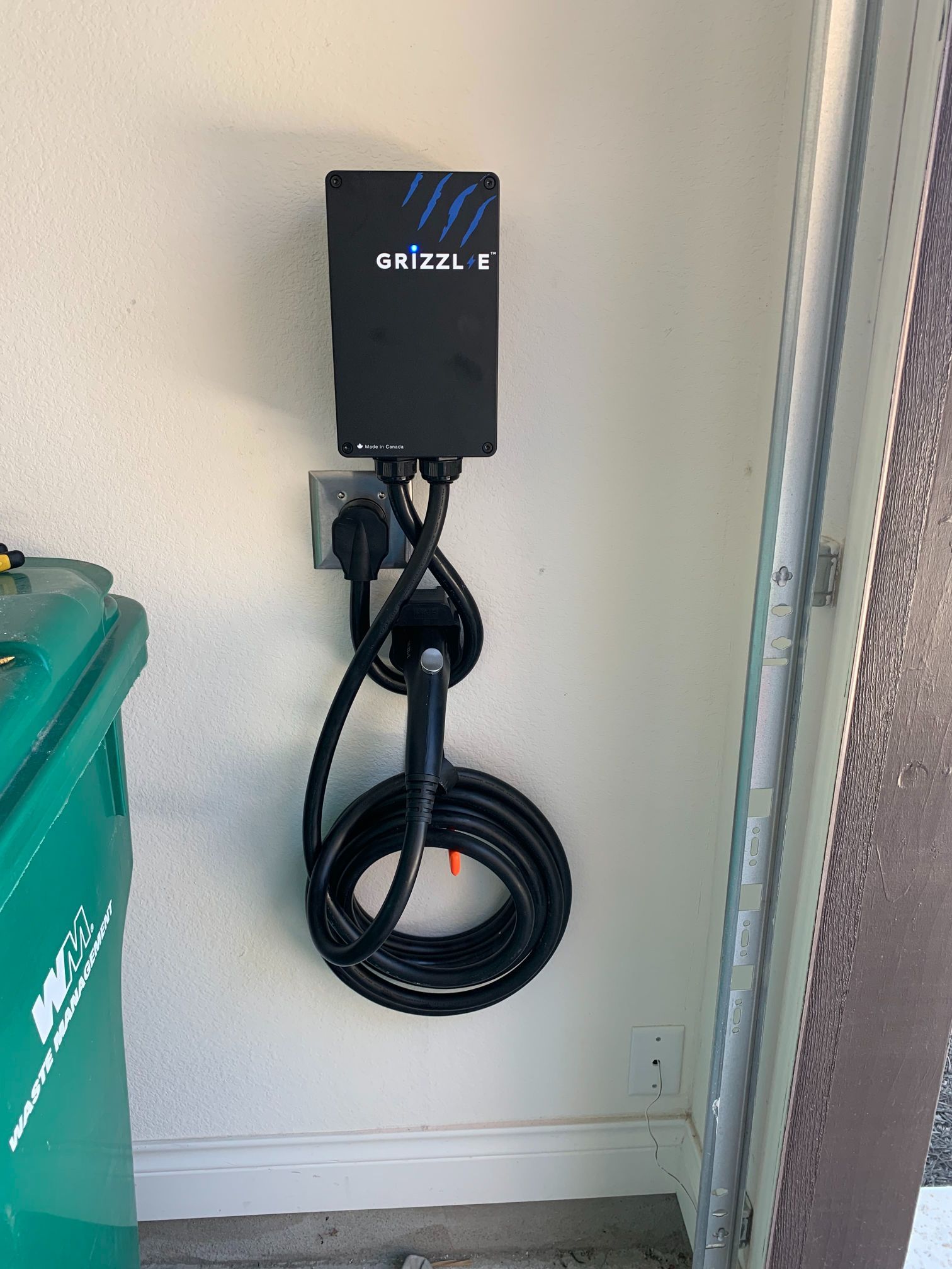 A black box with a hose attached to it is hanging on a wall next to a green trash can.