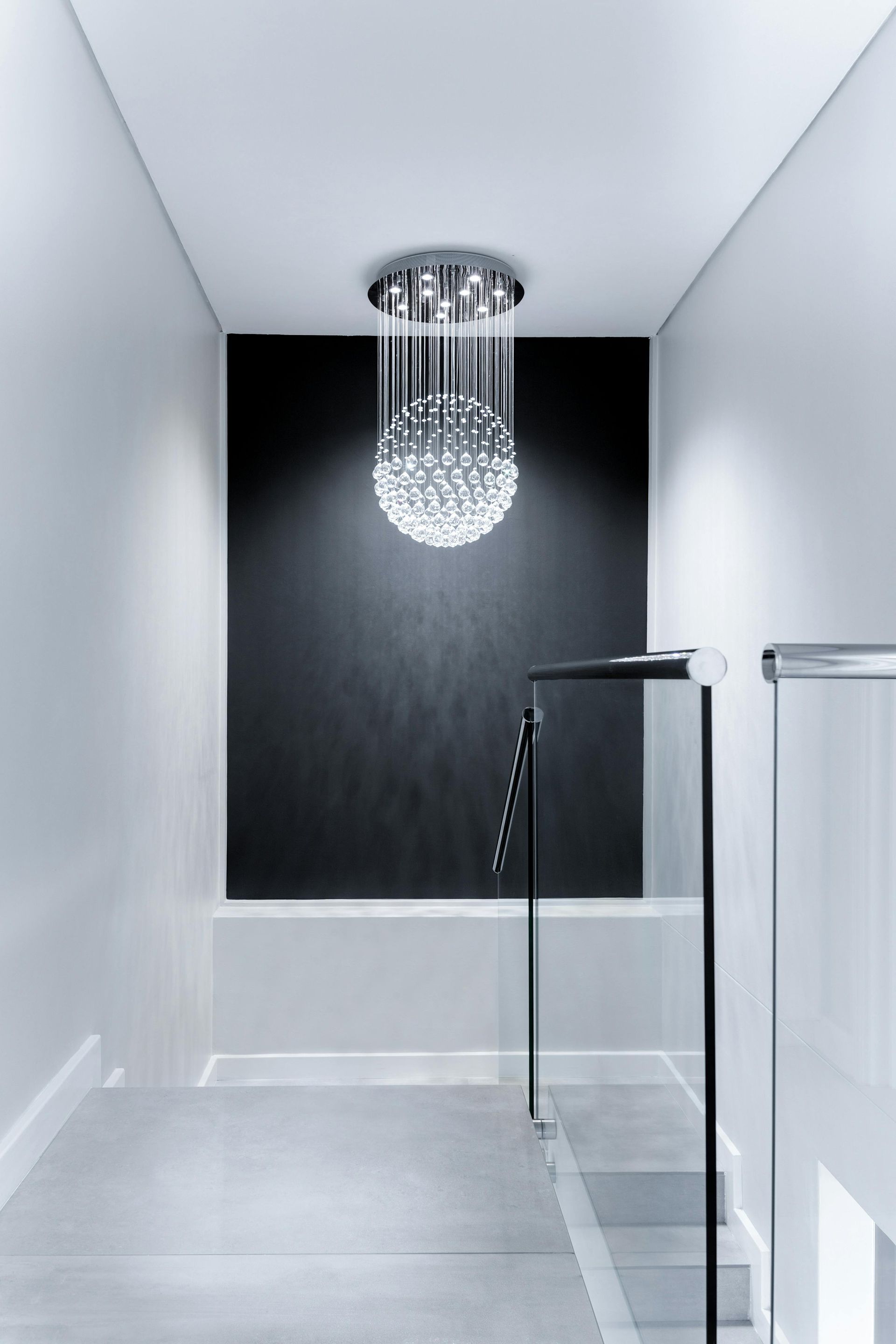 Narrow staircase with a modern chandelier centered on a black wall; glass railing.