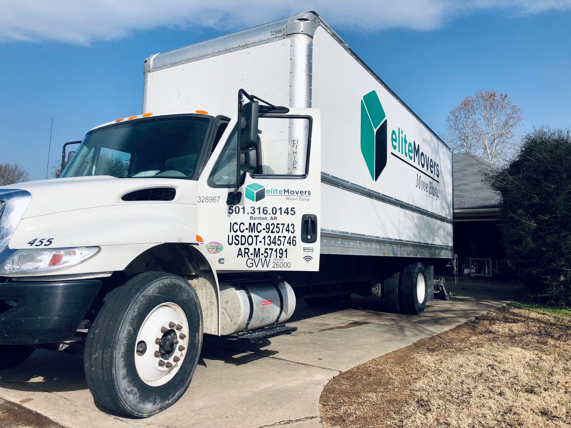 White moving truck parked in front of a house. The truck has the company logo, and details on it. Blue sky overhead.