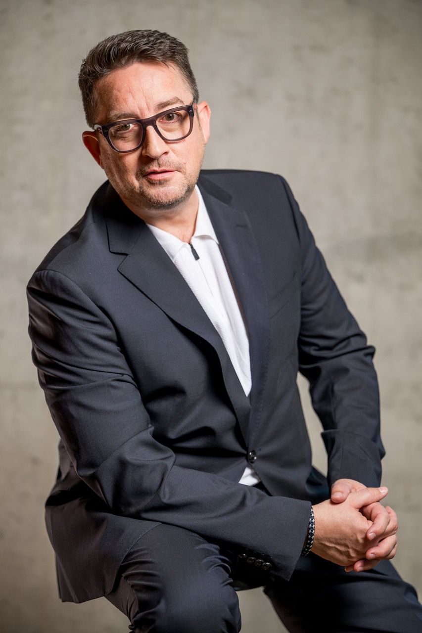 Mann in dunklem Anzug und Brille sitzt vor einem grauen Studiohintergrund, die Hände verschränkt.