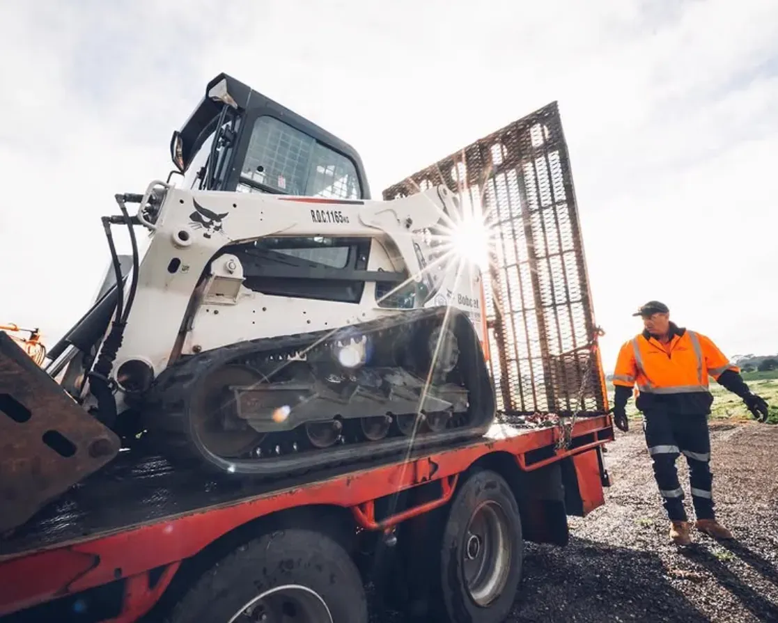 Bobcat track loader on a red truck bed, worker in orange safety vest walks alongside — Highlands Water Cartage in Glenquarry, NSW
