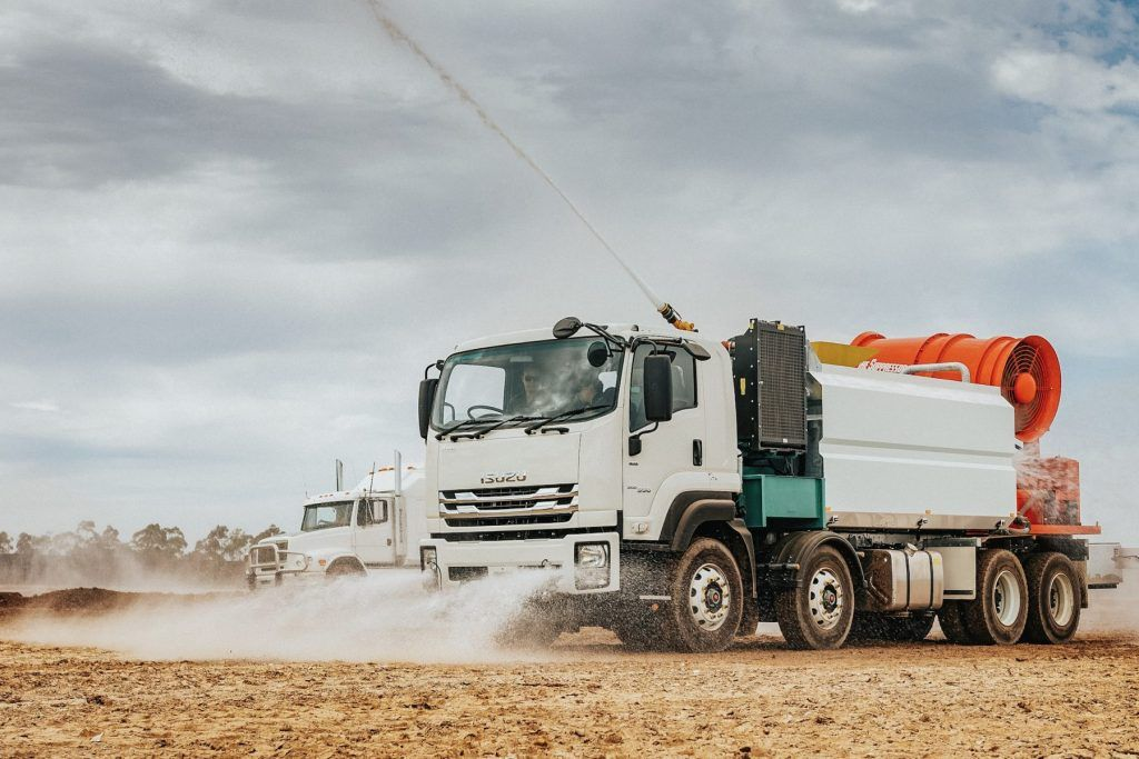 Water truck spraying dust suppressant on a dirt road. Bright sky, two trucks, spraying water — Highlands Water Cartage in Glenquarry, NSW