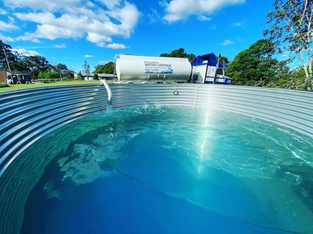 A large, corrugated metal water tank filled with blue water; a white water tank and blue equipment in the background — Highlands Water Cartage in Glenquarry, NSW